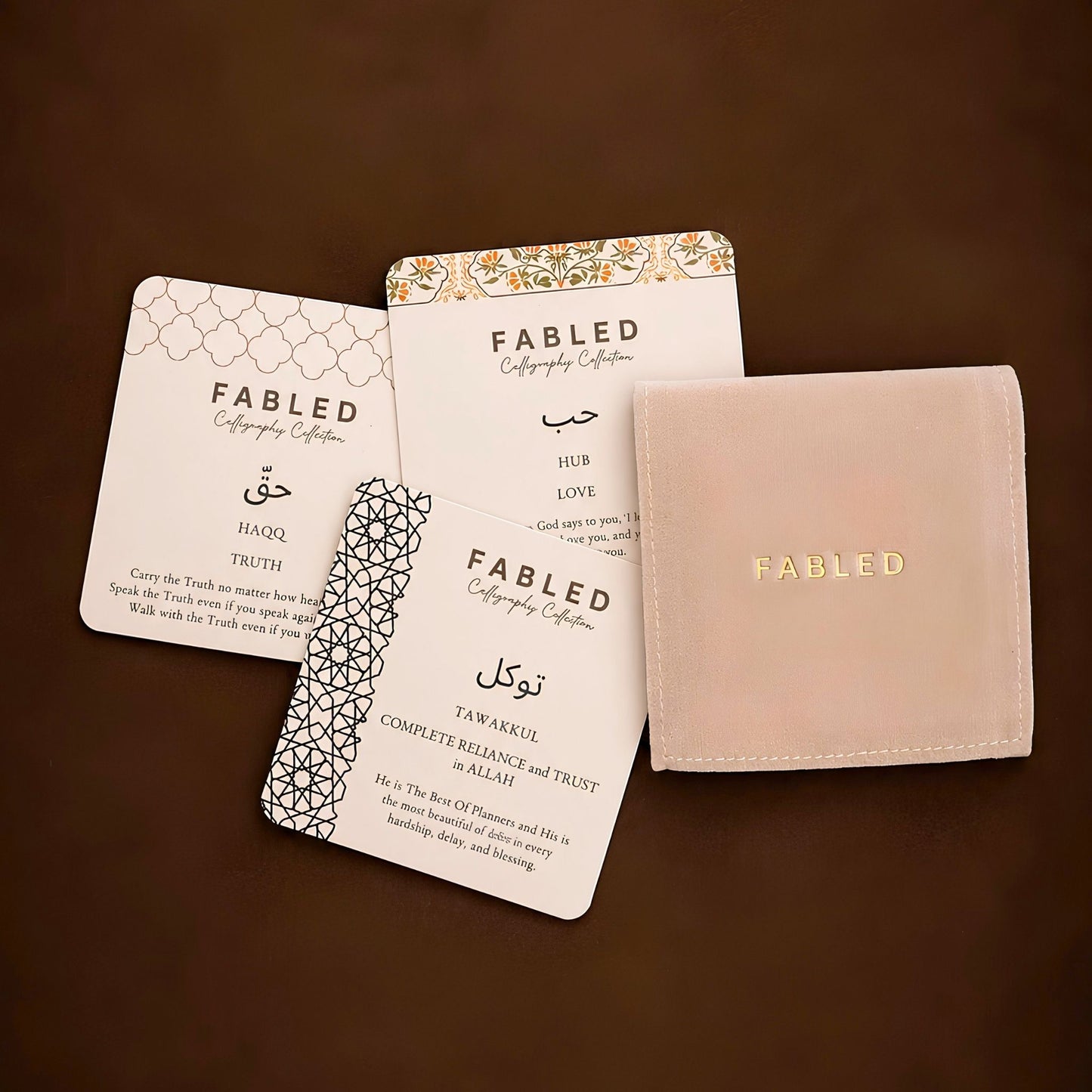 A coordinated set of Fabled calligraphy cards and a matching small pouch on a dark brown background. The set includes white cards with black text and geometric patterns, a patterned card partially tucked under another, and a pale blush pouch with gold 'FABLED' lettering.