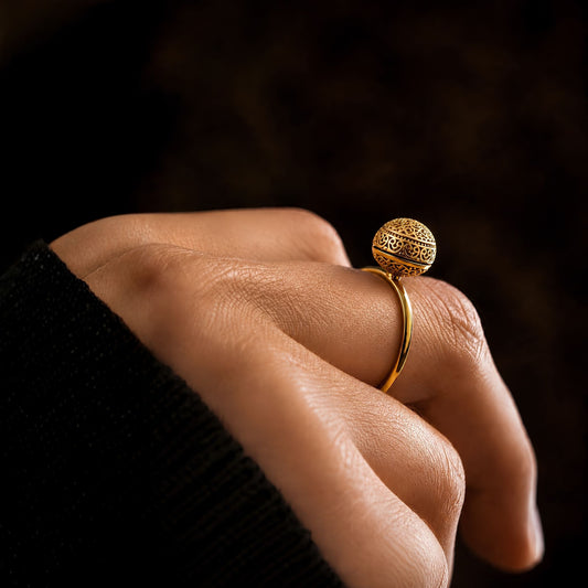 Gold ring with a detailed and layered floral motif draped over the surface of a minimalist spherical solitaire with a lovely antique finish on a finger against a dark background.