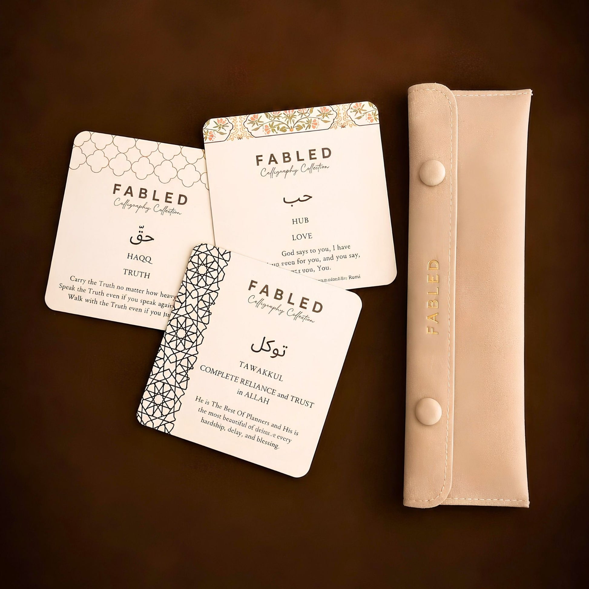A set of Fabled Calligraphy Collection wedding cards in cream with gold and black patterns, displayed next to a matching beige bracelet pouch on a dark brown background.