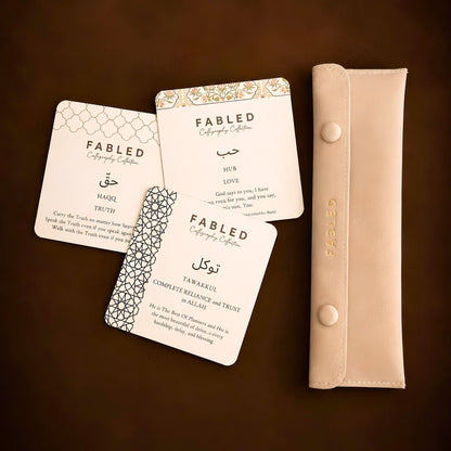 A set of Fabled Calligraphy Collection wedding cards in cream with gold and black patterns, displayed next to a matching beige bracelet pouch on a dark brown background.