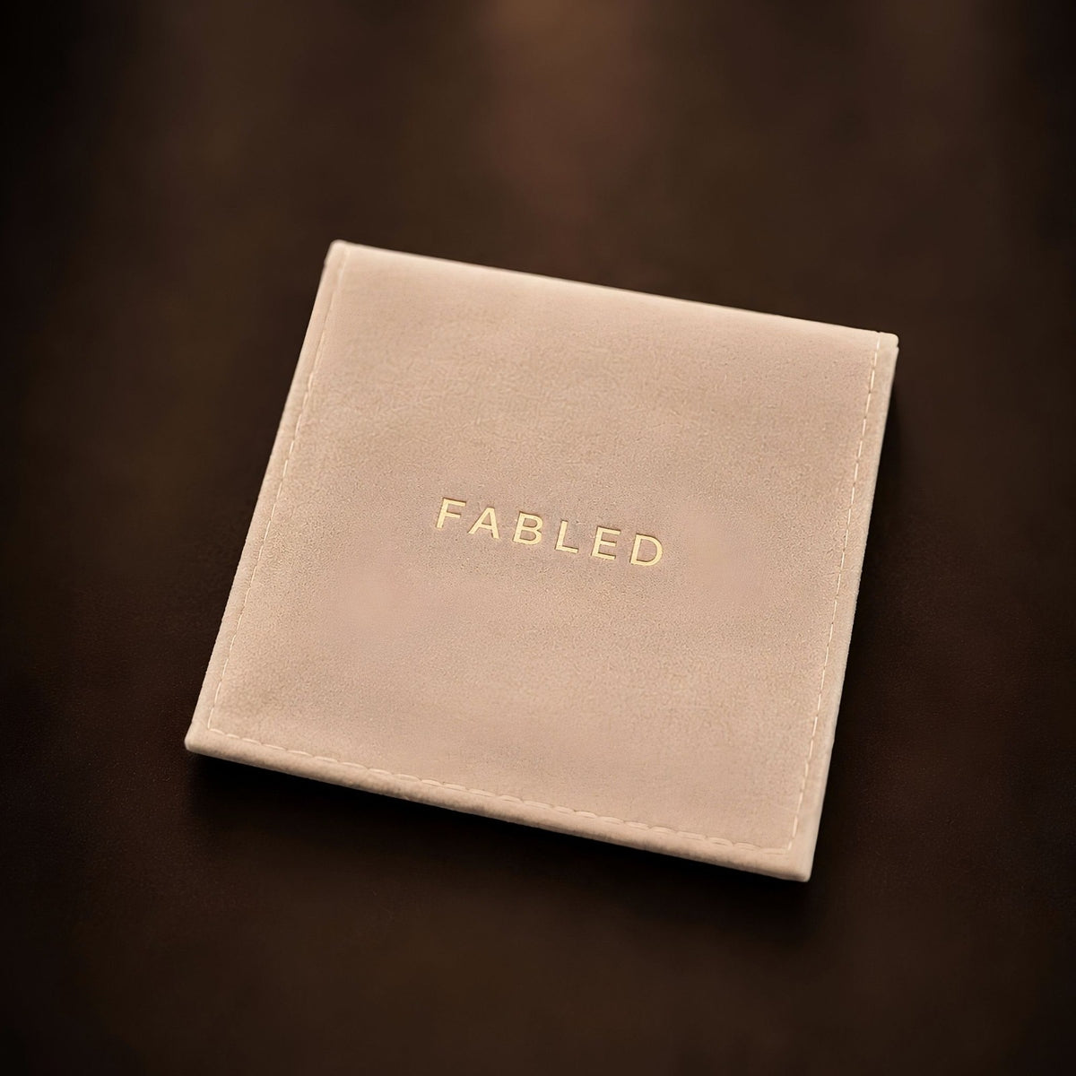 A square cream-colored pouch or with the brand name 'FABLED' embossed in gold lettering, resting on a dark brown background.