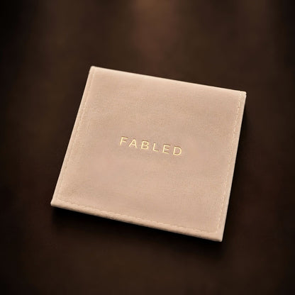 A square cream-colored pouch or with the brand name 'FABLED' embossed in gold lettering, resting on a dark brown background.