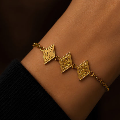 A gold plated chain bracelet with three diamond-shaped charms featuring traditional geometric and arabic calligraphy saying Maktub shown worn on a person's wrist.