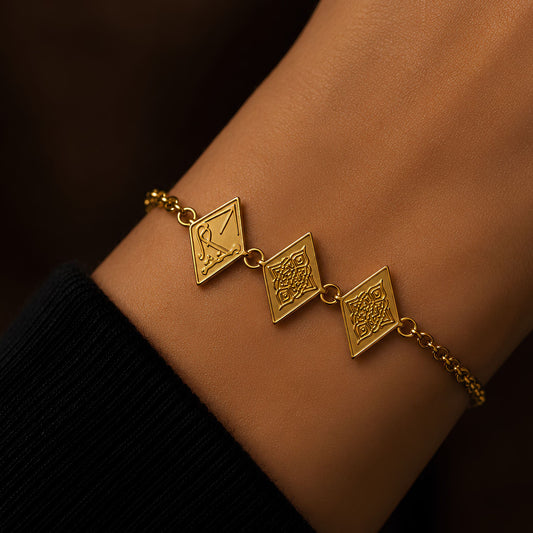 A gold plated chain bracelet with three diamond-shaped charms featuring traditional geometric and arabic calligraphy saying Maktub shown worn on a person's wrist.