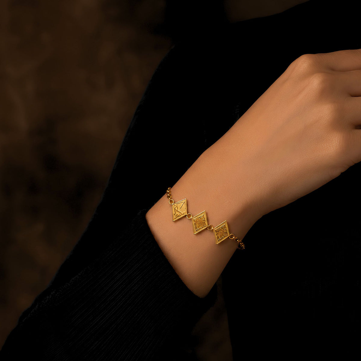 A delicate gold plated chain bracelet with three textured diamond charms worn on a person's wrist, shown against a dark black ribbed sweater and a dark brown background.