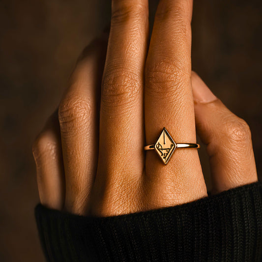 A close-up of a hand wearing gold plated Maktub ring featuring Arabic calligraphy by FABLED set against a dark, warm brown background.