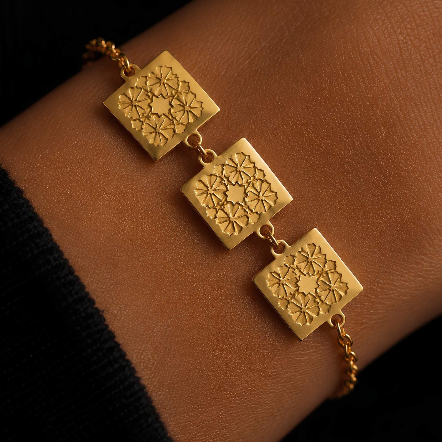Tapestry inspired stylized traditional floral rosette motif on a minimalist 3-charm gold plated bracelet, shown worn on a wrist against a warm brown skin background.