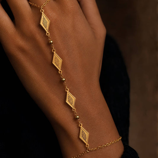 A gold hand chain bracelet with four dangling diamond-shaped charms engraved with traditional tapestry motif for a beautiful blend of Southeast Asian and Middle Eastern design and small bead accents, shown worn on a person's wrist and hand against a dark background.