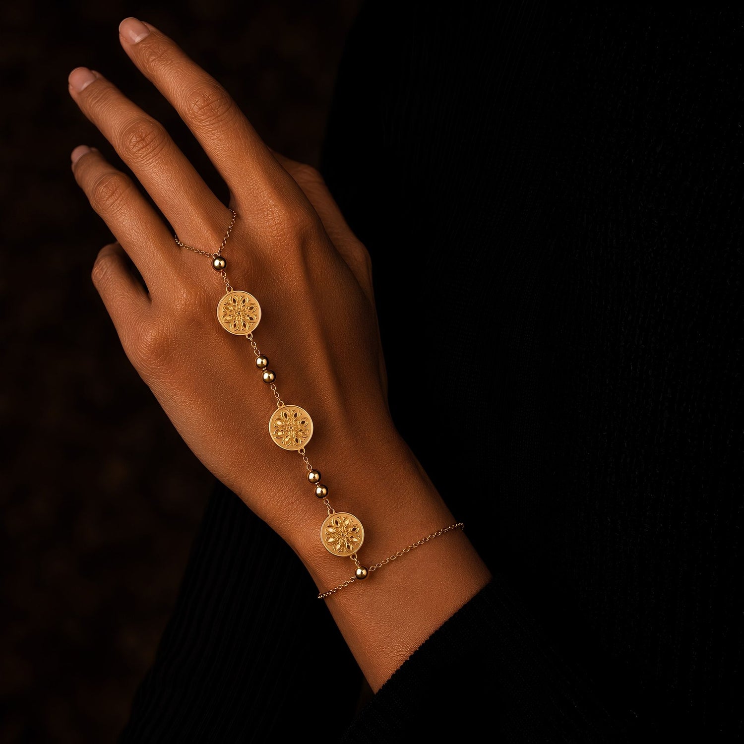 Gold hand chain bracelet with 8-petaled rosettes engraved onto a strand of coin-inspired pendants for a unique interpretation on a classic hand chain design.  on a wrist against a dark background. 