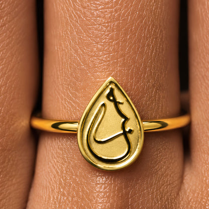 Gold plated ring with a teardrop-shaped emblem featuring Arabic calligraphy Love, shown worn on a finger.