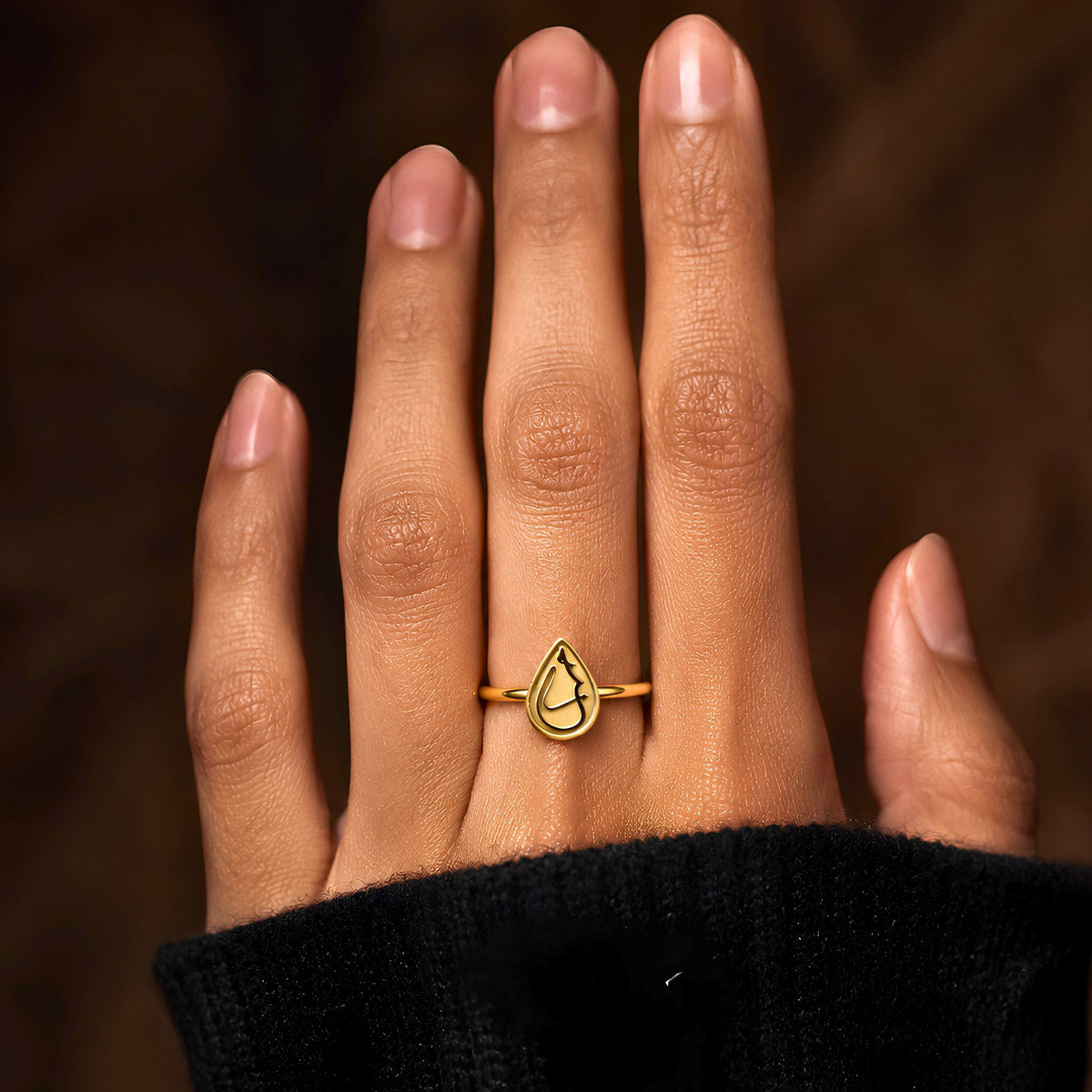 A gold plated ring with a teardrop-shaped outline and arabic calligraphy saying Love, worn on a person's finger. The hand is framed against a dark, warm brown background, and the person is wearing a black knit sleeve.