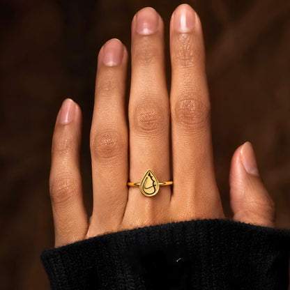 A gold plated ring with a teardrop-shaped outline and arabic calligraphy saying Love, worn on a person's finger. The hand is framed against a dark, warm brown background, and the person is wearing a black knit sleeve.
