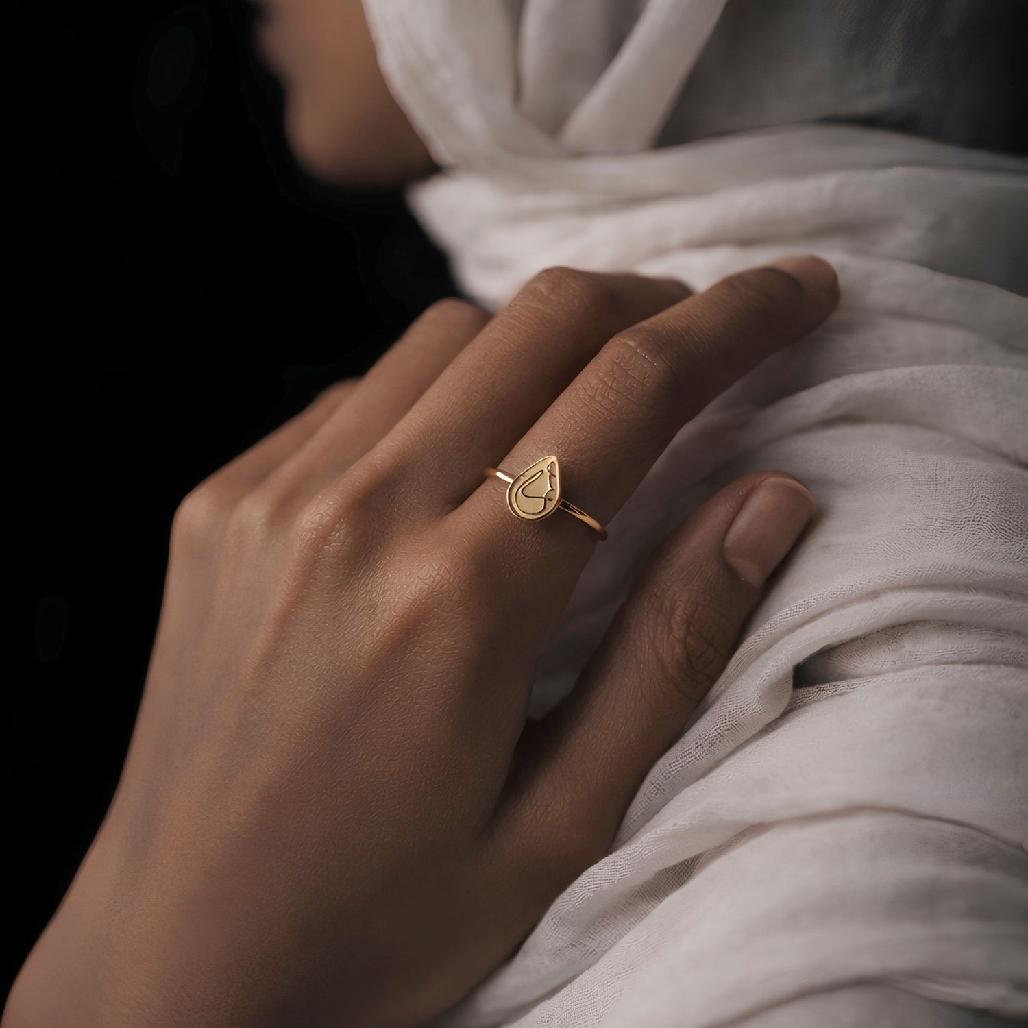 A delicate gold ring with a teardrop-shaped gold plated ring saying love worn on a finger, shown on a model draped in soft white fabric against a dark background.