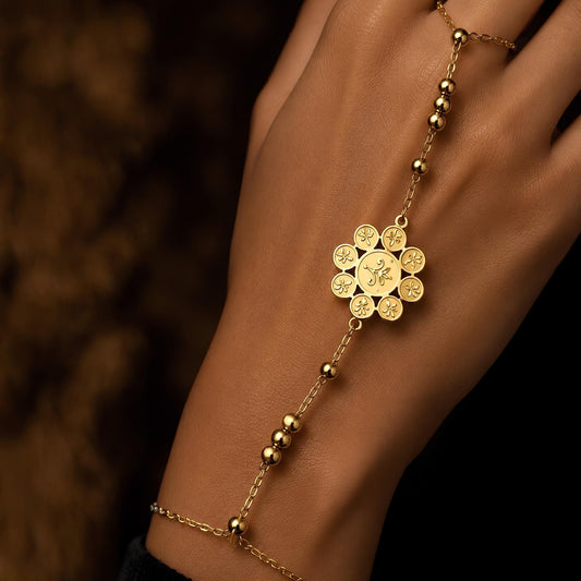 A close-up of a person's wrist and hand wearing a delicate gold hand chain bracelet with a stylized rosette motif sketched on the parts and petals of a classic floral pendant. 