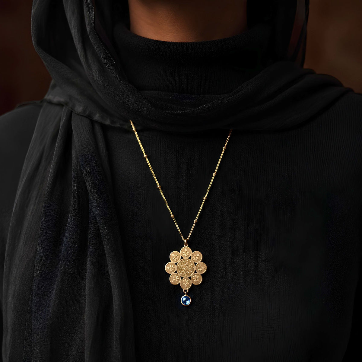 Gold necklace with a stylized rosette motif sketched on the parts and petals of a classic floral pendant with a royal sapphire blue dew drop gem on a black outfit.