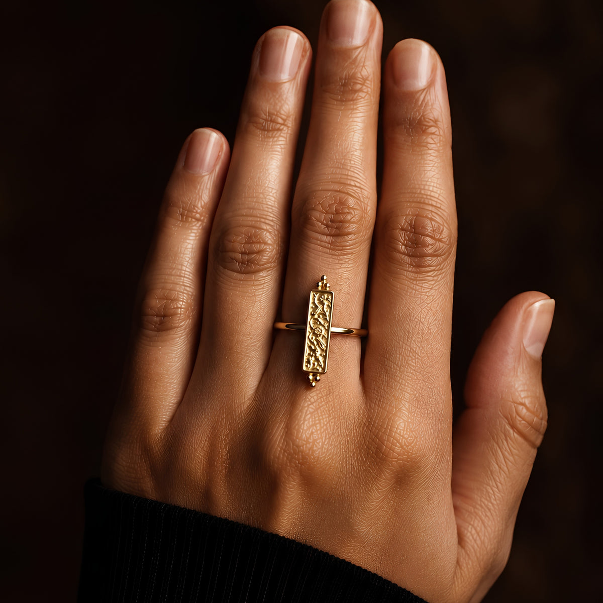 A close-up of a hand wearing a gold plated ring with a textured vertical bar showcasing a vintage floral motif in an antique finish, framed within a thin panel and crowned by stacked bead bookends, on a dark background.
